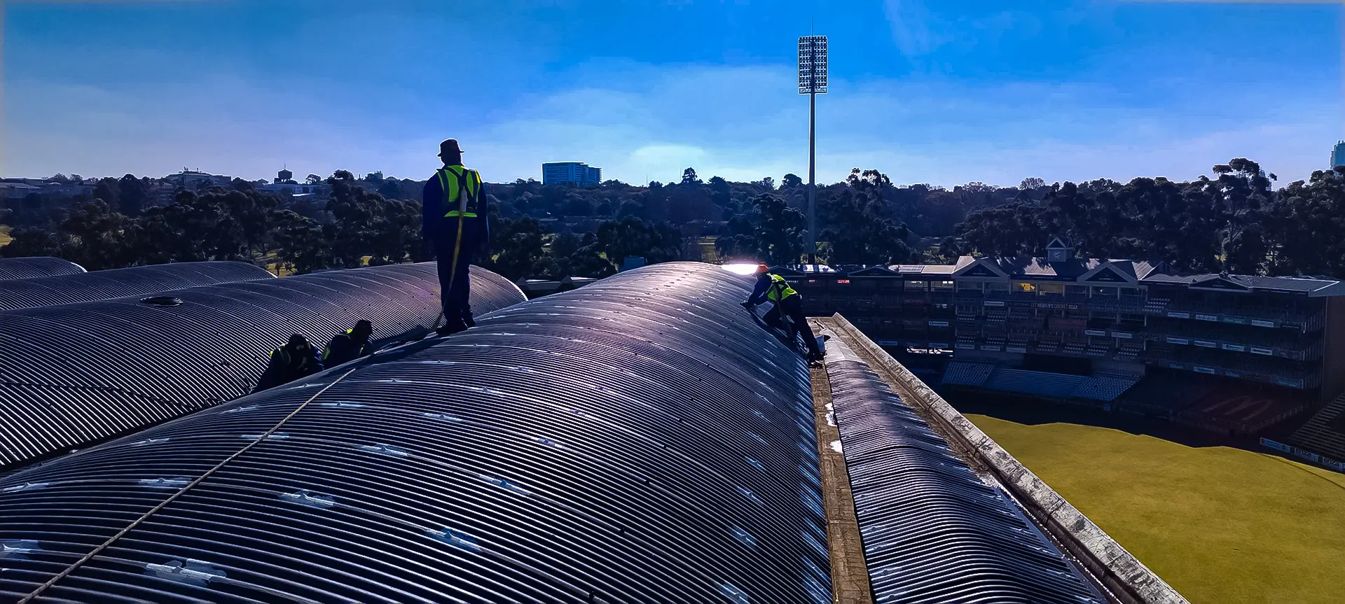 Local roof leak repair experts near me diligently inspect a vast corrugated stadium roof on a sunny day, ensuring peak condition.